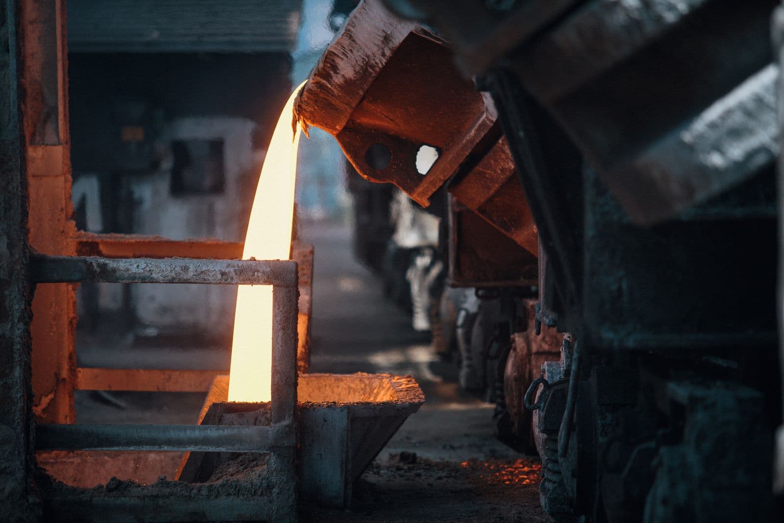 Stream of molten metal pouring from an industrial foundry ladle, with sparks scattering across the pour floor.